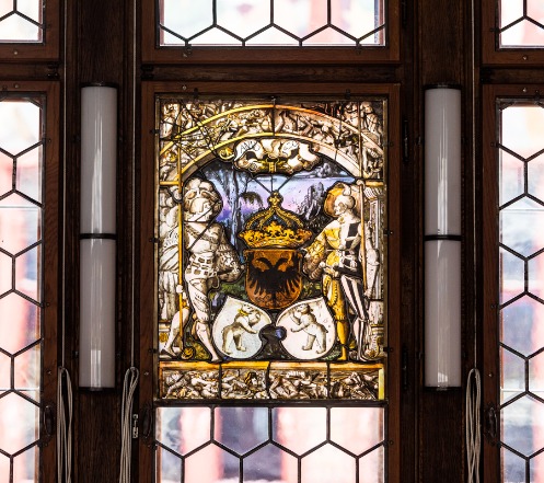 Eine Glasmalerei des Rathauses. Im Zentrum der Malerei befindet sich ein Wappen mit einem doppelköpfigen Adler. Über dem Wappen ist eine Krone abgebildet. Zu beiden Seiten des Wappens sind zwei Männer mit Speeren gemalt. Unter dem Wappen sind zwei weitere Wappen abgebildet mit je einem Bären darin.